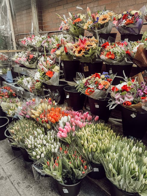 A display of freshly arranged flower bouquets in black buckets, featuring a variety of colorful flowers including pink, white, orange, and purple tulips, carnations, and lilies. The arrangements are w