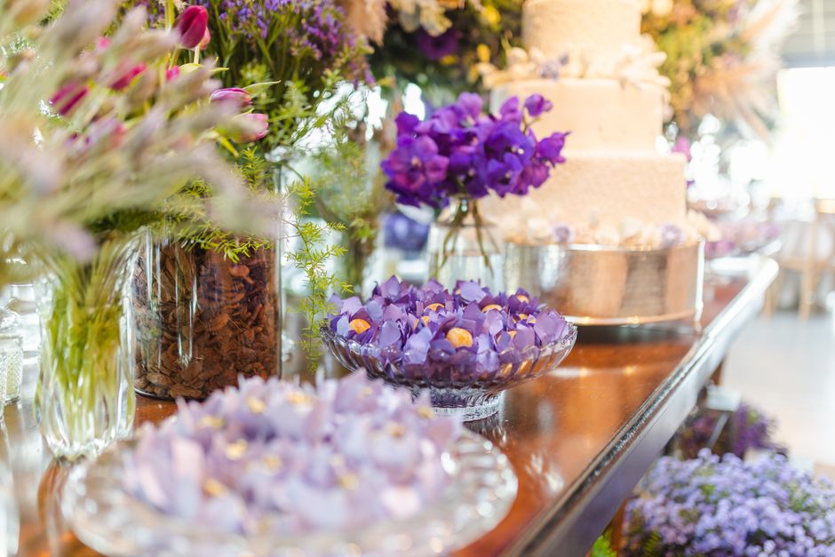 A variety of fresh floral arrangements displayed on a polished wooden table, featuring purple and lavender flowers including violets and orchids, arranged in clear glass bowls, vases, and wrapped bouq