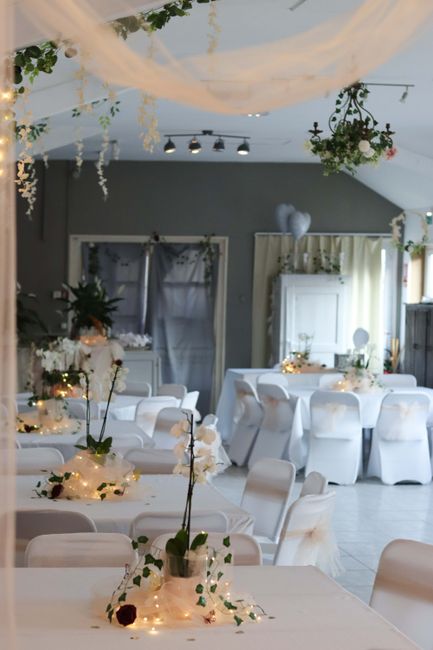 A wedding venue decorated with elegant floral arrangements and soft lighting, featuring tables draped in white tablecloths with floral centrepieces and fairy lights. The arrangement in focus includes 