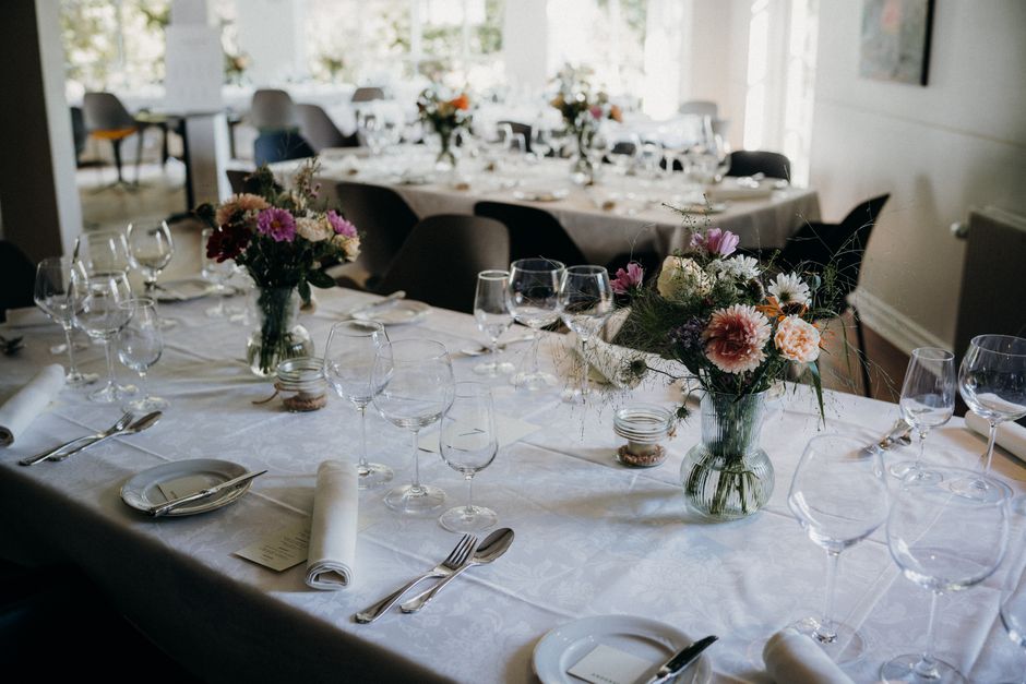A floral arrangement featuring a mix of pink, white, and purple flowers, including carnations, daisies, and chrysanthemums, displayed in a clear glass vase on a white linen-covered table. The table se